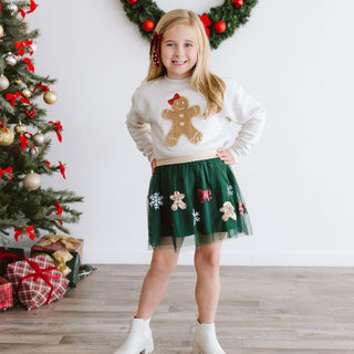 Sweet Wink Gingerbread Sequin Patch Christmas Sweatshirt - Natural
Young girl in a Christmas-themed outfit with a sweater and skirt, standing in front of a decorated Christmas tree.