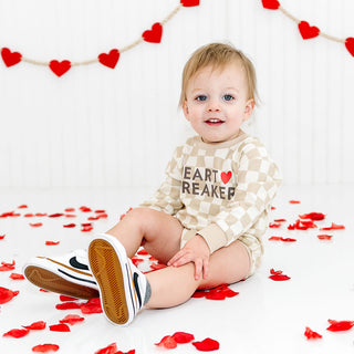 Sweet Wink Heartbreaker Checkered Valentine's Day Long Sleeve Romper - Natural Child wearing a checkered shirt with text, surrounded by red heart decorations and petals on a white background