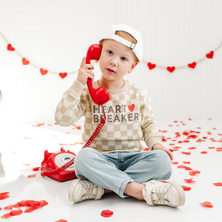 Sweet Wink Heartbreaker Checkered Valentine's Day Sweatshirt - Natural
Child holding a red phone with hearts around, sitting on a white floor with heart decorations.