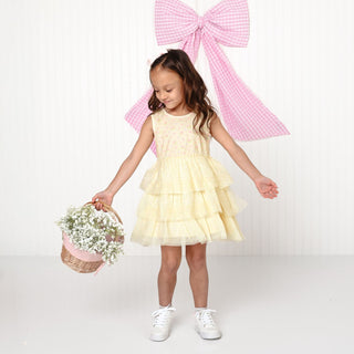 Sweet Wink Lemon Floral Tank Tutu Dress
Young girl in a yellow dress holding flowers with a large pink bow in the background