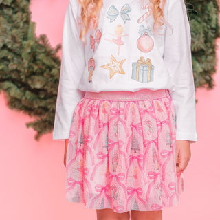 Sweet Wink Nutcracker Cutie Christmas Long Sleeve Shirt - White Person wearing a white long-sleeve shirt with festive designs and a pink skirt with bow pattern against a pink background.