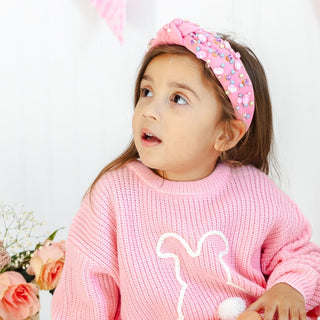 Sweet Wink Pastel Gemstone Headband
Young girl wearing a pink headband and sweater with a white design, sitting in front of flowers.