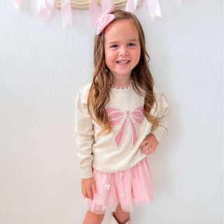 Sweet Wink Pearl Bow Patch Sweatshirt - Natural
Young girl wearing a cream sweater with a pink bow and a pink skirt, standing against a white background.