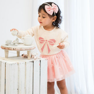Sweet Wink Pearl Bow Patch Sweatshirt - Natural
Young girl in a pink and white outfit with a bow, playing with a tea set.