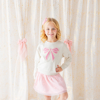 Sweet Wink Pearl Bow Patch Sweatshirt - Natural
Young girl wearing a white sweater with a pink bow and a pink skirt, standing in front of a curtain.