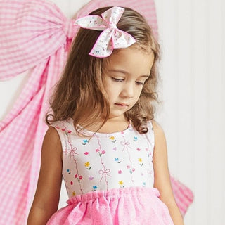 Sweet Wink Peony Floral Bow Clip
Sweet Wink Peony Floral Tank Tutu Dress
Young girl in a pink and white dress with a large bow in her hair against a pink background