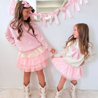 Sweet Wink Pretty Pearl Petal Tutu
Two young girls in matching pink and white outfits with bows, standing in a room with decorative ribbons.