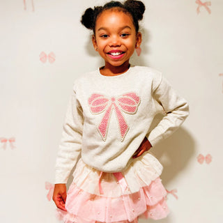 Sweet Wink Pretty Pearl Petal Tutu
Young girl wearing a sweater with a pink bow design against a light background