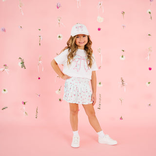 Sweet Wink Sky Blue Floral Bow Patch Short Sleeve T-Shirt - White
Young girl in a white t-shirt with a bow and floral shorts standing on a pink background with decorative elements.