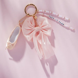 Little Words Project Bag Charm - Take It Slow
A pink ballet shoe charm keychain with a large pink satin bow, a gold key ring, and a beaded strap that reads 'TAKE IT SLOW', displayed on a soft pink background.