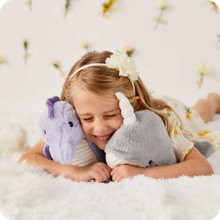 Seahorse Warmies
Young girl hugging two plush toys, one purple and one gray, on a soft surface.