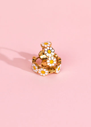 Pierced Co. Huggies - Daisy
A gold-toned ring decorated with multiple white daisy flowers with yellow centers, set against a solid light pink background.