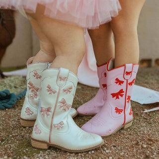 Wild & Rae cowgirl boots with white leather, pink bow accents, pointed toe, and Western-style stitching on the shaft.