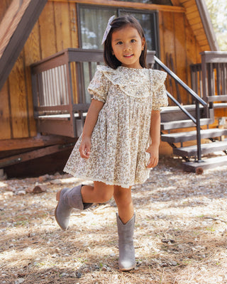Rylee & Cru Winnie Dress - Winter Garden
A young girl wearing a cream-colored floral print dress with ruffled shoulders and gray cowboy boots, standing outdoors in front of a wooden cabin.