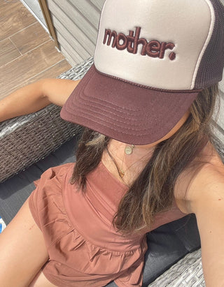 "Mother." Embroidered Trucker Hat - Brown + Tan
Person wearing a cap with 'mother' text sitting on a chair.