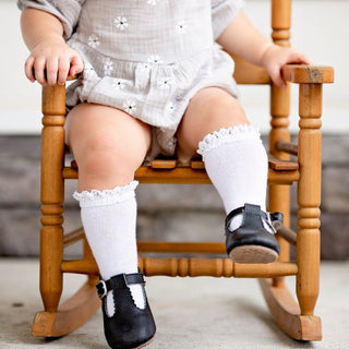 Little Stocking Co Lace Top Knee High Socks - White
Child sitting on a wooden rocking chair wearing white socks and black shoes.