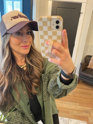 "Mother." Embroidered Trucker Hat - Brown + Tan
Woman taking a mirror selfie wearing a cap and green jacket, holding a phone with a checkered case.