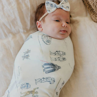 Copper Pearl Star Wars Tatooine™ Swaddle Blanket
Baby wearing a white outfit with black illustrations and a bow headband, lying on a soft surface.