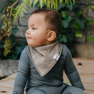 Copper Pearl Star Wars Tatooine™ Bandana Bib Set
Baby wearing a gray bib with a pattern, sitting outdoors with greenery in the background