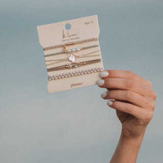 K'lani "Glimmer" Soft Pinks with Pearl and Gold Accent Hair Tie Bracelets
Hand holding a package of gold and silver hair ties with 'kelani' branding on a gray background