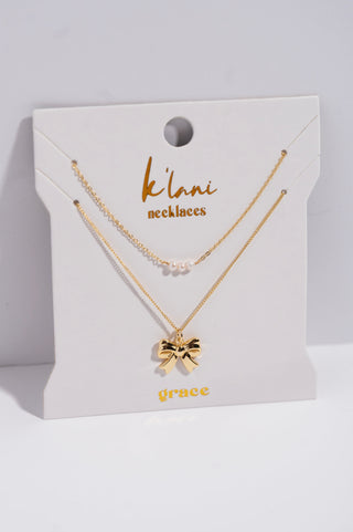K'lani "Grace" Gold Bow with Pearl Accent Necklace Set
A gold necklace set on a white display card, featuring a delicate chain with a small gold bow pendant and a second chain with tiny pearl-like beads. The card is branded 'k'lani necklaces' and 'grace'.