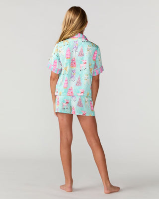 Sunshine Society Kids Silky Short Sleeve Short Set - Prancer
A woman stands barefoot against a light gray studio background, viewed from the back, wearing a matching short-sleeve pajama top and shorts set in mint green with a pink and multicolor holiday tree pattern.
