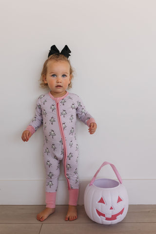 In My Jammers Ballerina Ghost Zipper Romper Child in a pink floral onesie with a black bow, standing next to a white pumpkin bucket on a light wooden floor.