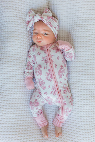 In My Jammers Blush Garden Zipper Romper
Newborn baby in a pink floral onesie and headband lying on a textured surface.