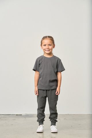 Little Bipsy Waffle Top - Shadow
Young girl wearing a gray t-shirt and pants standing against a plain white wall.