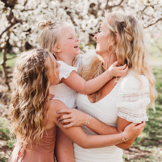 K'lani "Mother / Daughter" Pink with Pearls and Gold Hearts Shareable Hair Tie Bracelets
Two women and a young girl hugging in a park with cherry blossom trees in the background