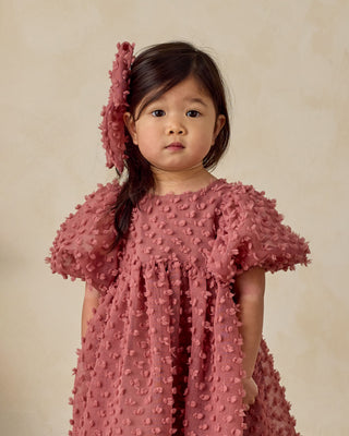 Noralee Everly Bow - Raspberry
Young girl wearing a pink textured dress and matching headband against a beige background