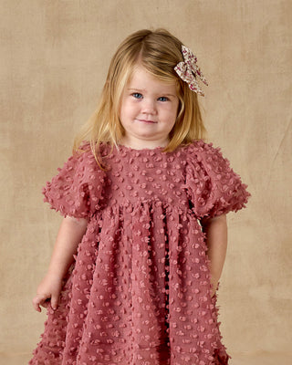 Noralee Luna Dress - Raspberry
Young girl wearing a pink dress with a textured pattern against a beige background