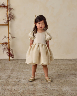 Noralee Sofia Dress - Ecru
Young girl in a beige dress standing in a room with a wooden ladder and dried plants.