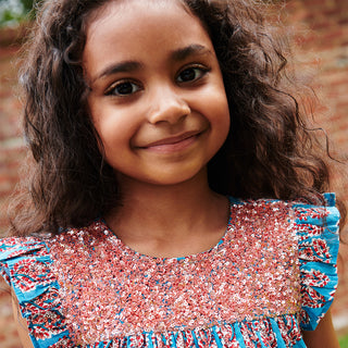 Pink Chicken Girls Fiona Ruffle Dress - Blue Paisley Young girl wearing a colorful sequined dress with a blurred brick wall background