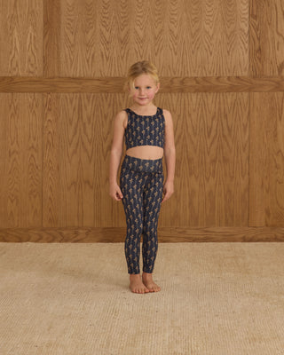 Play by Rylee & Cru Swift Sports Bra - Vintage Bloom Play by Rylee & Cru Basic Legging - Vintage Bloom Young girl wearing a navy patterned outfit standing against a wooden paneled wall.