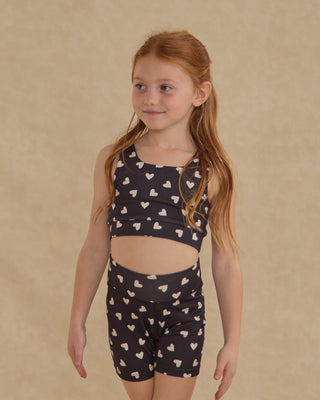 Play by Rylee & Cru Swift Sports Bra - Hearts
Young girl wearing a black tank top and shorts with heart patterns on a beige background