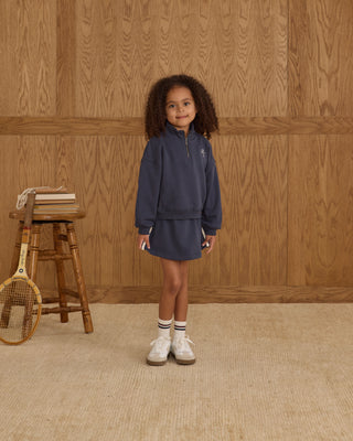 Play by Rylee & Cru Half Zip Sweatshirt - Navy Play by Rylee & Cru Sports Skirt - Navy Child wearing a navy tracksuit standing in front of a wooden paneled wall.