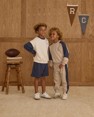Play by Rylee & Cru Paneled Sweatpant - Stone Two children in matching outfits standing in a room with wooden walls and a stool with a football.