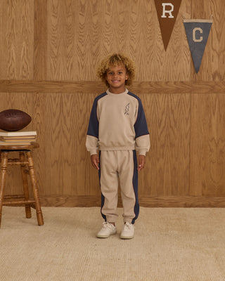 Play by Rylee & Cru Paneled Sweatshirt - Stone Child wearing a beige and navy tracksuit standing in front of a wooden wall with sports-themed decor.