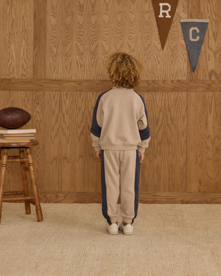 Play by Rylee & Cru Paneled Sweatshirt - Stone Child wearing a beige and navy tracksuit standing in front of a wooden wall with decorative elements.
