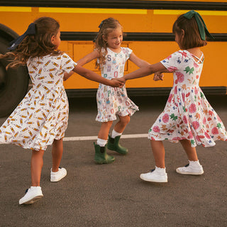 Posh Peanut Posh Pencils Ruffled Twirl Dress Three young girls in patterned dresses holding hands in front of a yellow school bus.
