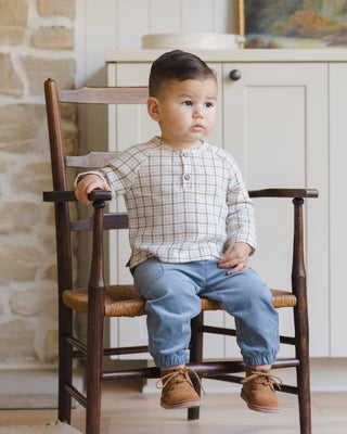 Quincy Mae Zion Shirt - Blue Plaid
Child sitting on a wooden chair in a room with stone walls and a white cabinet.