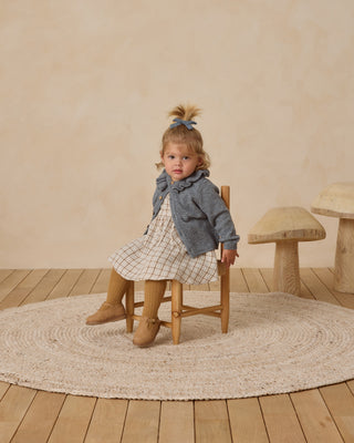 Quincy Mae Simone Dress - Blue Plaid
Young girl sitting on a wooden chair in a room with a wooden floor and beige walls.