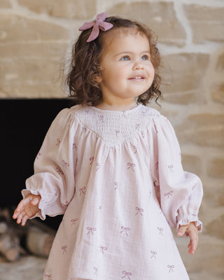 Quincy Mae V Smocked Dress - Bows
Child wearing a light pink dress with bow patterns in front of a stone wall.