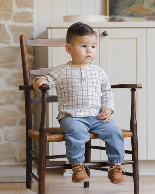 Quincy Mae Zion Shirt - Blue Plaid
Child sitting on a wooden chair in a room with stone walls and a white cabinet.