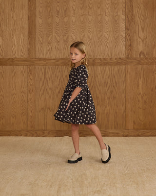 Rylee & Cru Rose Dress - Hearts
Young girl in a black dress with white patterns standing against a wooden paneled wall.