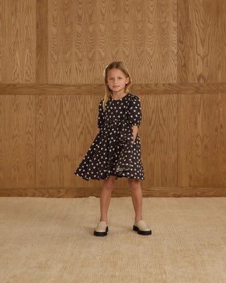 Rylee & Cru Rose Dress - Hearts
Young girl wearing a black dress with white patterns against a wooden paneled wall.