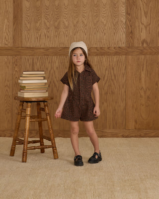 Rylee & Cru Utility Romper - Leopard
Young girl in a brown dress standing next to a wooden stool with books against a wooden paneled wall.