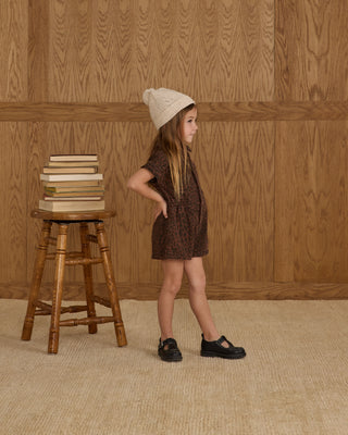 Rylee & Cru Utility Romper - Leopard
Young girl in a brown dress and beige hat standing next to a wooden stool with books against a wooden paneled wall.