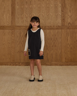 Rylee & Cru V-Neck Overall Dress - Washed Black
Young girl wearing a black dress with a white shirt underneath against a wooden paneled wall.